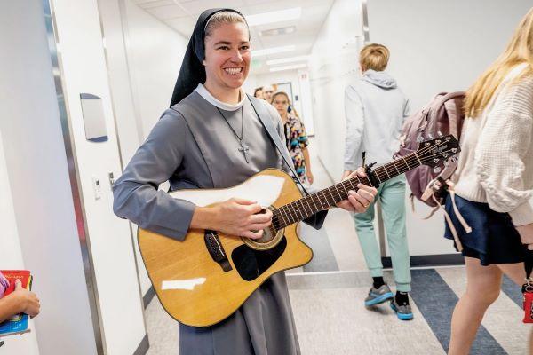 Sister M. Xavier Schulze smiles while playing guitar in a school hallway